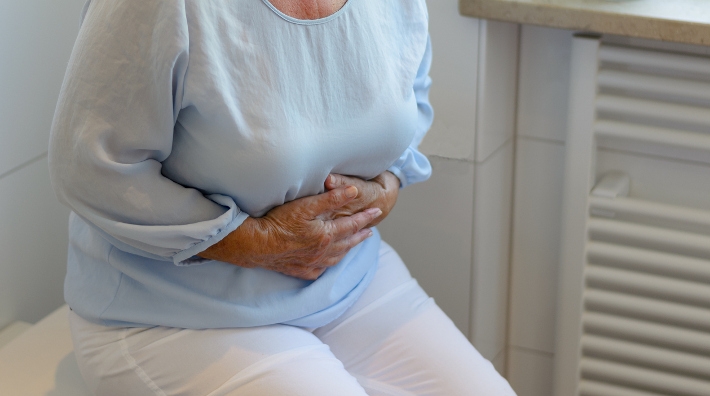 Mid section of senior woman wearing white trousers and light blue blouse sitting on toilet holding stomach