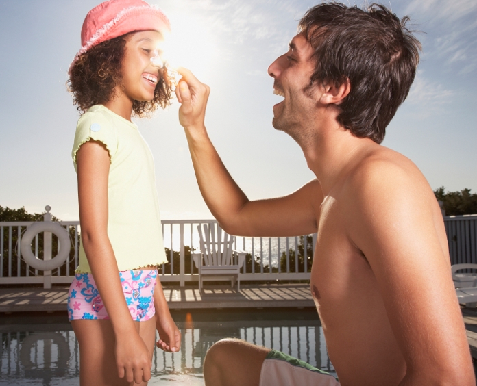 father applying sunscreen to his young daughter's face at pool